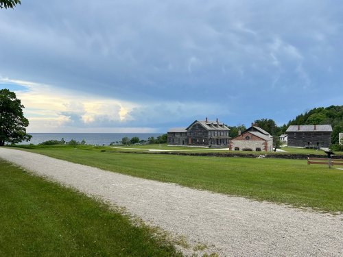 A gravel path leads to historic buildings by the water under a cloudy sky, with green grass in the foreground.