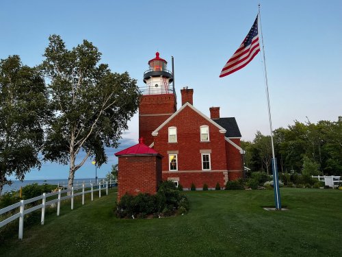 A red brick lighthouse with a white lantern, surrounded by greenery and a flagpole displaying the American flag.