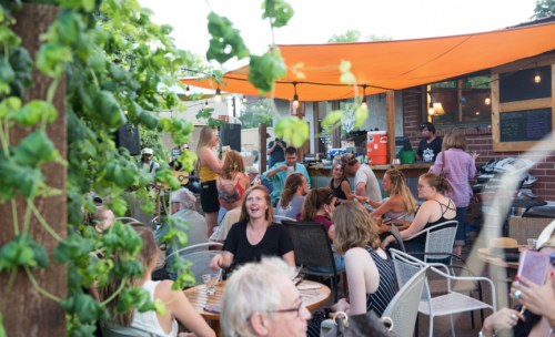 A lively outdoor gathering at a café with people socializing under an orange canopy, surrounded by greenery.