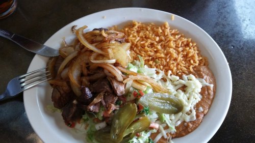 A plate of Mexican food featuring grilled meat, sautéed onions, rice, refried beans, lettuce, tomatoes, and jalapeños.