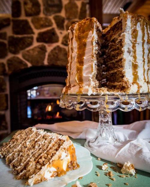 A layered cake with caramel drizzle, partially sliced, on a glass cake stand, with a cozy fireplace in the background.