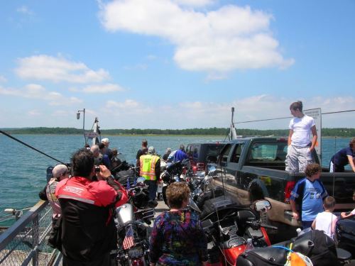 A busy ferry with motorcycles and trucks, passengers waiting by the water on a sunny day.