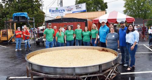 A large cooking pan filled with food, surrounded by a group of people in green shirts at an outdoor event.
