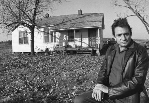 A man in a leather jacket sits in front of a small, weathered house surrounded by bare trees and fallen leaves.