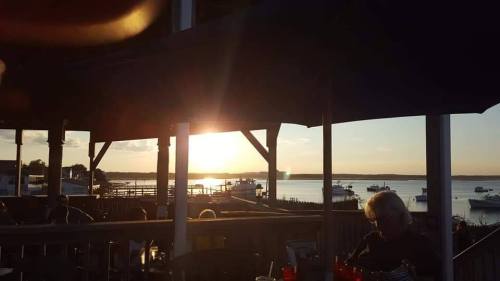 Sunset over a calm bay, with boats in the water and silhouettes of umbrellas and people enjoying the view.