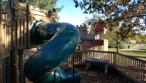 A green slide curves down from a wooden play structure in a sunny park, with trees and a playground in the background.