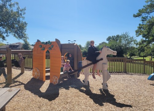 A playground scene with children playing on a wooden horse and a carriage structure under a clear blue sky.