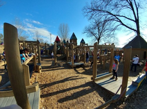 A vibrant playground filled with children playing, featuring wooden structures and colorful play equipment under a clear blue sky.