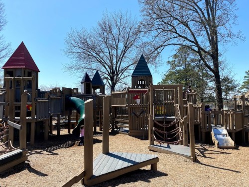 A wooden playground with various structures, slides, and climbing areas, surrounded by trees under a clear blue sky.