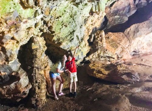 Two people explore a cave, posing next to rock formations and illuminated by soft lighting.