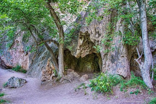 A rocky cave entrance surrounded by trees and greenery, with a dirt path leading towards it.