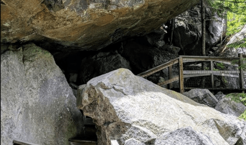 A rocky cave entrance with large boulders and a wooden walkway leading into the shaded area.