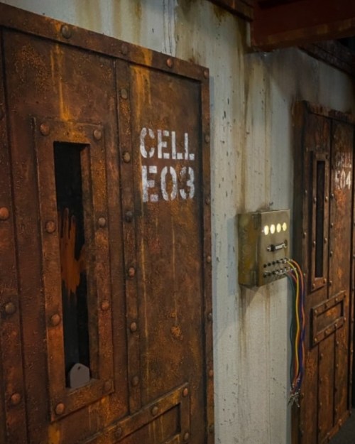 Rusty prison cell doors labeled "CELL E03" and "CELL E04," with a control panel featuring buttons and wires.