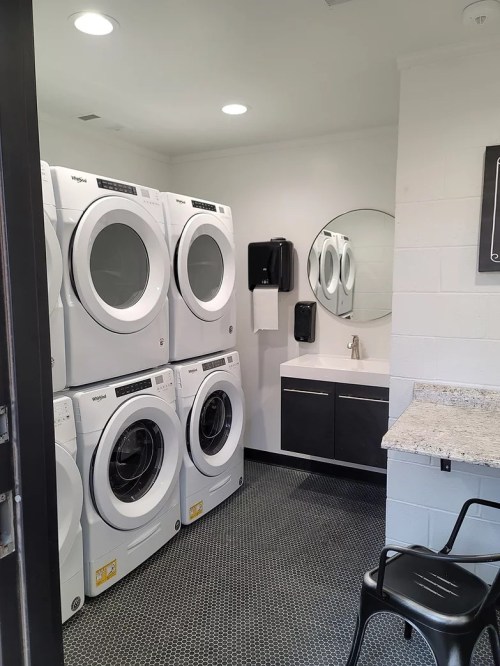 A modern laundry room featuring stacked washers and dryers, a sink, mirror, and a small table with a chair.