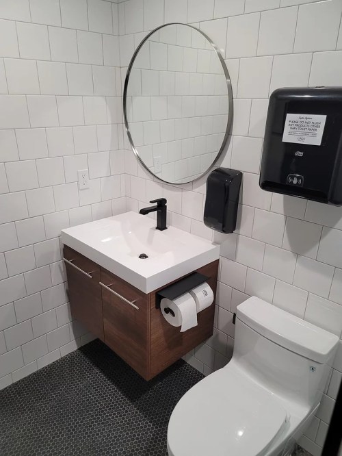 Modern bathroom with a round mirror, sink, wooden cabinet, toilet, and paper dispenser on tiled walls.