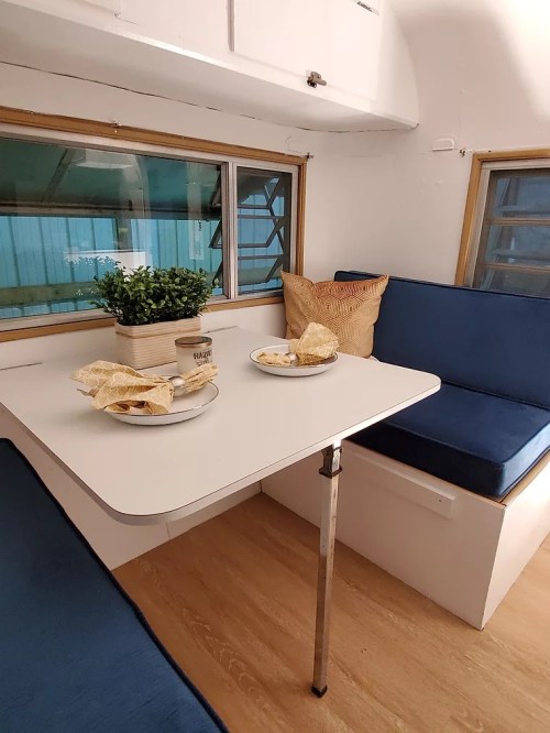 Cozy interior of a small camper with a table, blue seating, and a potted plant, creating a welcoming atmosphere.