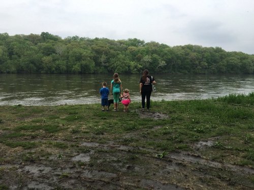 A group of children and an adult stand by a riverbank, looking out at the water and lush green trees in the background.