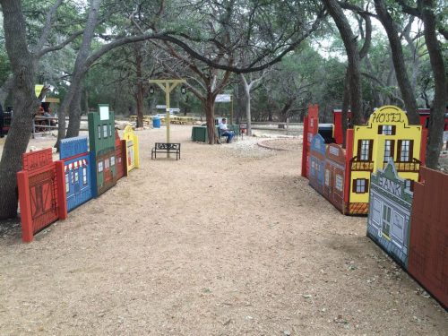 A pathway lined with colorful wooden buildings in a park, surrounded by trees and gravel.