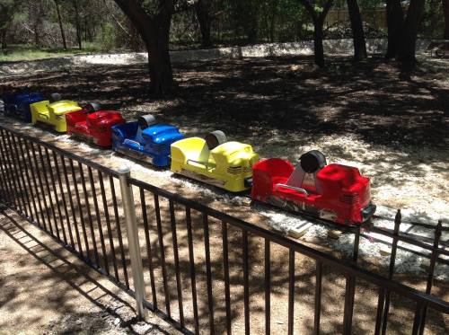A colorful miniature train on tracks, featuring red, blue, yellow, and green cars, set in a wooded area.