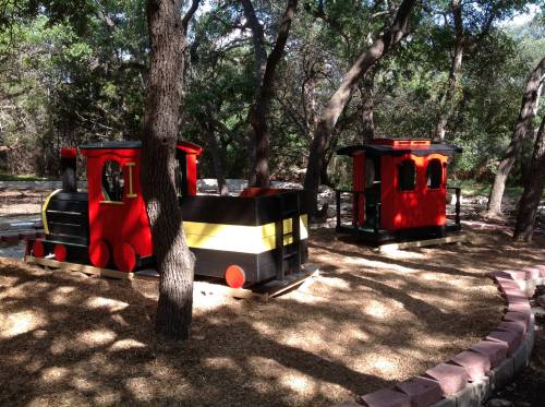 Colorful wooden train play structure surrounded by trees and mulch in a park setting.