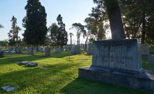 A serene cemetery scene with gravestones surrounded by trees and green grass, illuminated by soft morning light.
