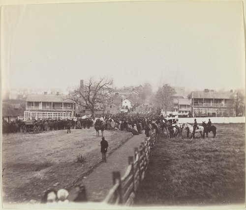 A historical black-and-white photo of a large crowd gathered near buildings, with horses and soldiers in the foreground.