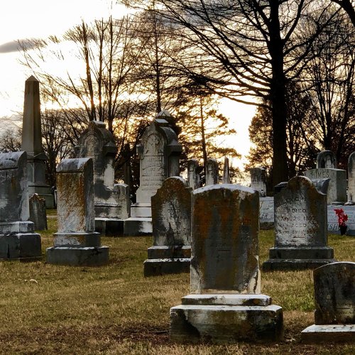 A cemetery at sunset, featuring weathered gravestones and bare trees against a cloudy sky.