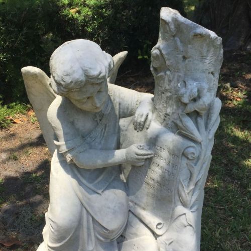 A marble angel statue kneeling beside a stone, with intricate details and a serene expression in a garden setting.