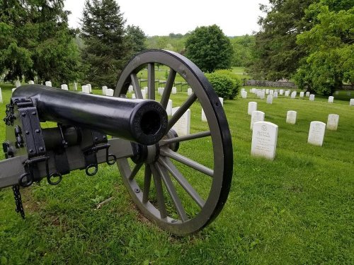 A historic cannon in the foreground with a cemetery and gravestones visible in the background, surrounded by greenery.