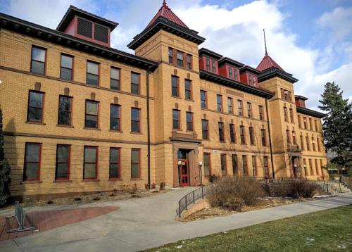Historic brick building with a red roof, large windows, and a landscaped entrance, set against a cloudy sky.