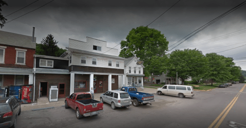 A small town street scene featuring several parked vehicles and a two-story building with a porch. Overcast sky.