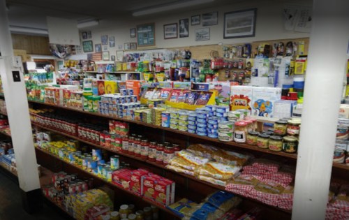 A well-stocked grocery store aisle filled with various canned goods, snacks, and packaged foods on wooden shelves.