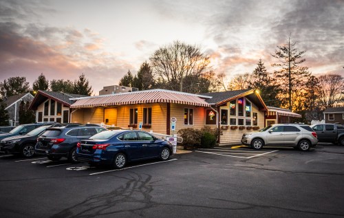 A cozy restaurant with a wooden exterior, surrounded by parked cars, at sunset with colorful lights glowing.