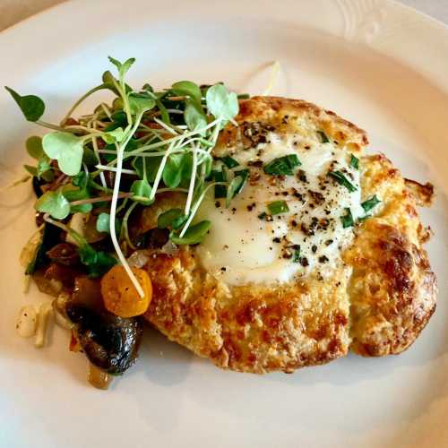 A golden-brown baked dish topped with an egg, herbs, and microgreens, served with sautéed vegetables on the side.