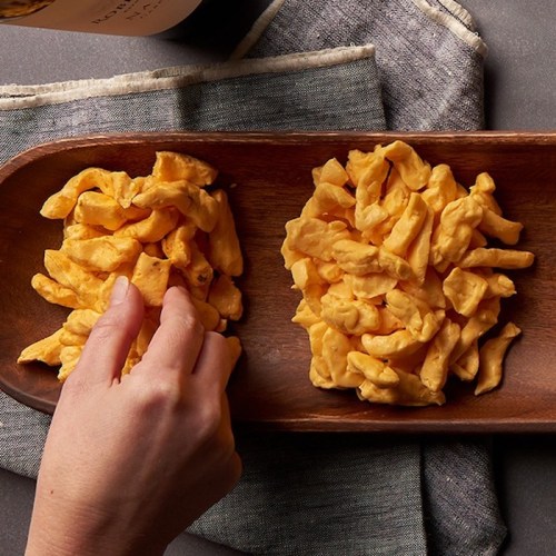 A hand reaching for a wooden platter filled with orange cheese puffs on a textured gray surface.