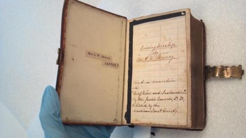 An open vintage book with handwritten notes, held by a gloved hand, showing a label and personal inscriptions.