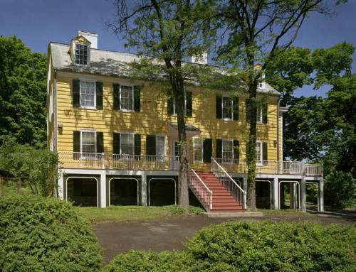 A large yellow house with green shutters, featuring a staircase and surrounded by trees and greenery.