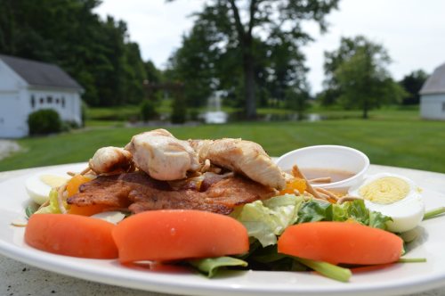 A colorful salad topped with grilled chicken, sliced tomatoes, hard-boiled eggs, and a side of dressing, set outdoors.