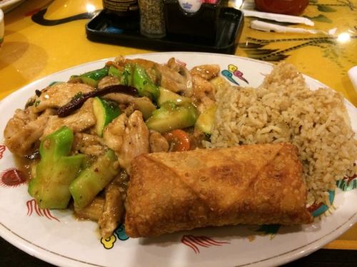 A plate of stir-fried chicken with vegetables, served with fried rice and a crispy egg roll on the side.