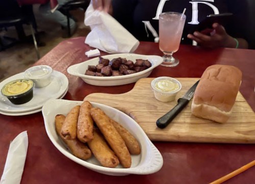 A table with a plate of fried cornmeal sticks, a serving of diced meat, a roll, and a drink.