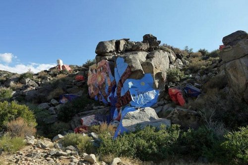 Colorful rock art on boulders amidst a rocky landscape with shrubs and blue sky in the background.