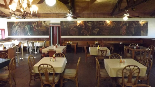 A spacious restaurant interior with wooden beams, tables set with white tablecloths, and large murals on the walls.