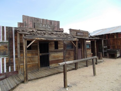 A rustic western-style building with a jail and sheriff's office, featuring wooden structures and a sandy path.