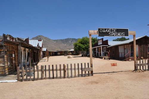 A dusty street in a ghost town with wooden buildings and a sign reading "Cuyamde Springs" under a clear blue sky.