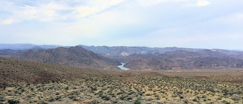 A panoramic view of rugged mountains and a winding river, surrounded by arid land and sparse vegetation under a cloudy sky.