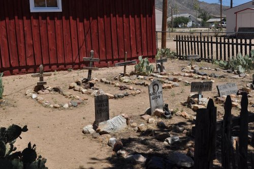 A small graveyard with wooden crosses and headstones, surrounded by rocks and cacti, near a red building.
