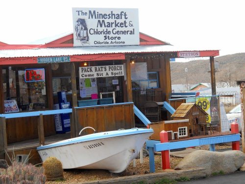 A rustic general store with a sign, porch seating, and a boat displayed outside in a small Arizona town.