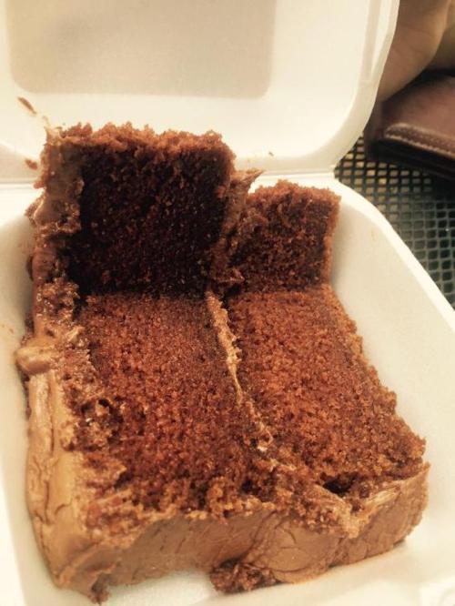 A slice of chocolate cake in a takeout container, showing moist layers and rich frosting.