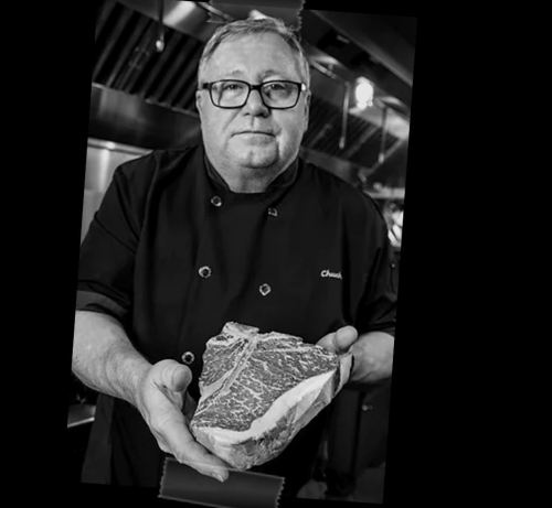 A chef in a black coat holds a large cut of meat, standing in a professional kitchen.