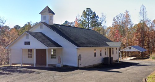 A white church building surrounded by trees with autumn foliage, featuring a steeple and a small covered area nearby.